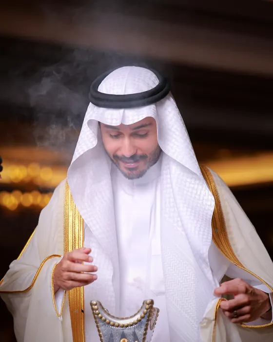 A host in a white thobe and bisht offering the incense burner to his guests